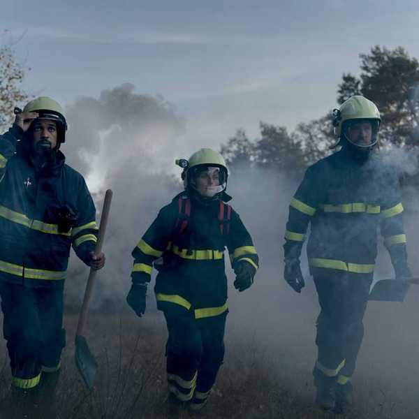 Three firefighters in turnout gear — woman in the center, flanked by two men carrying shovels in smoky air