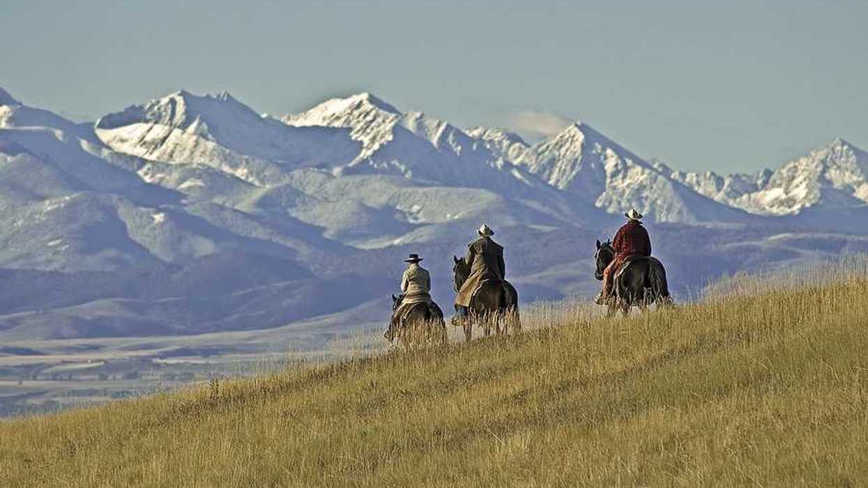 Three people on horses riding over a grassy hill toward mountains
