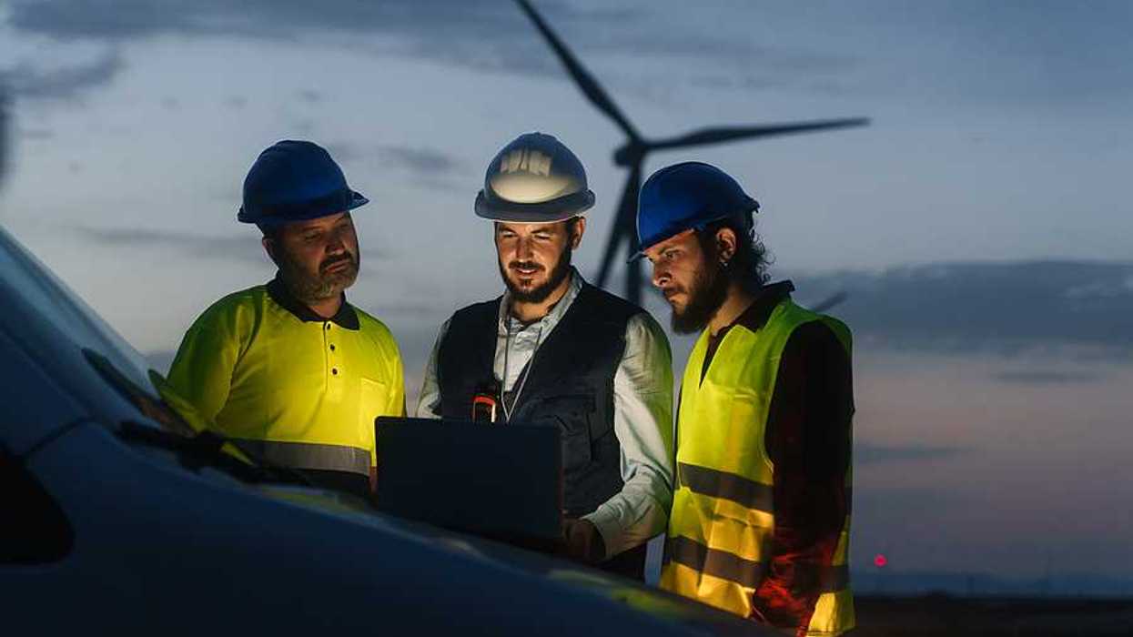 Three renewable energy workers looking at a laptop with a wind turbine in the background