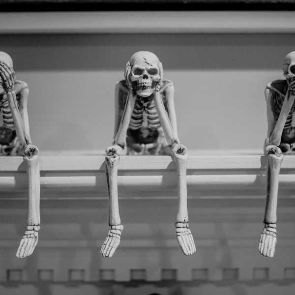 Three seated skeletons in a row covering eyes, ears, and mouth, respectively.