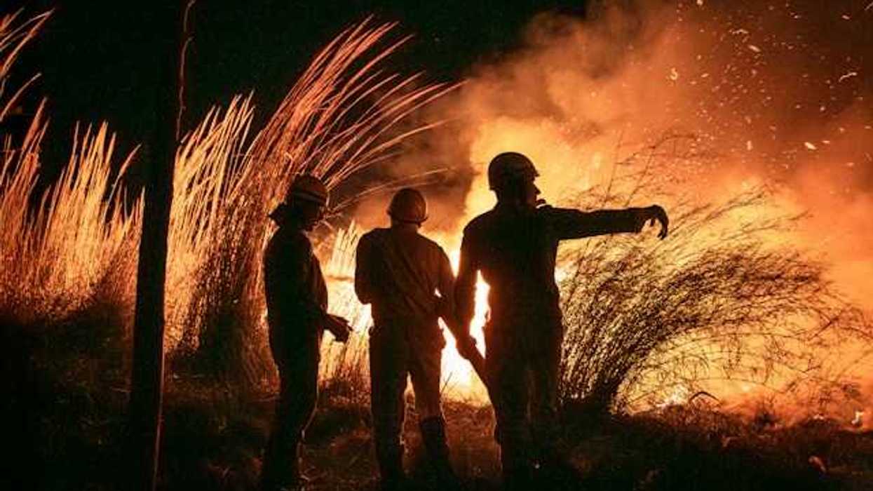 Three silhouetted figures watch a large fire burn in a field