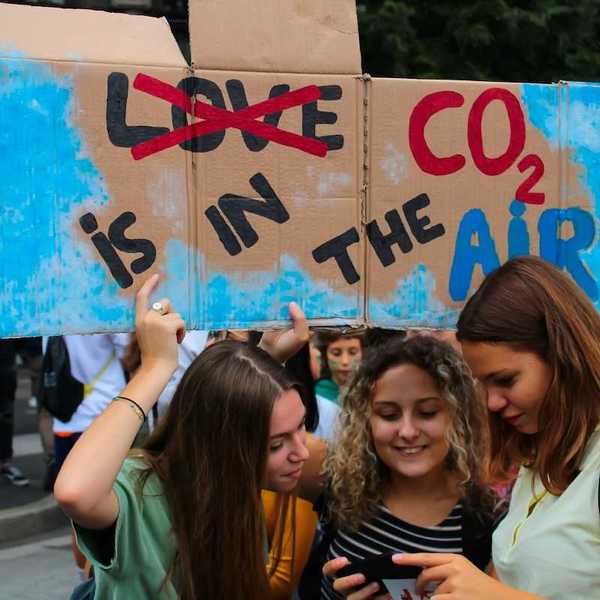 Three young women huddling over a phone or tablet, bearing a handmade sign reading "LOVE CO2 IS IN THE AIR" with the word love crossed out.