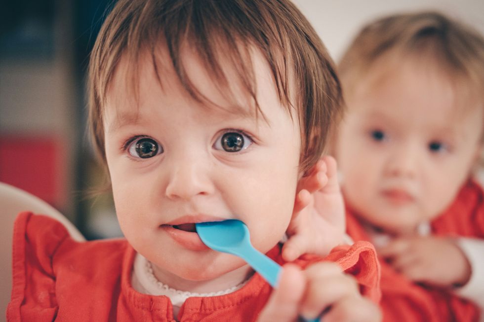 toddler putting a blue spoon in mouth with another toddler in background.