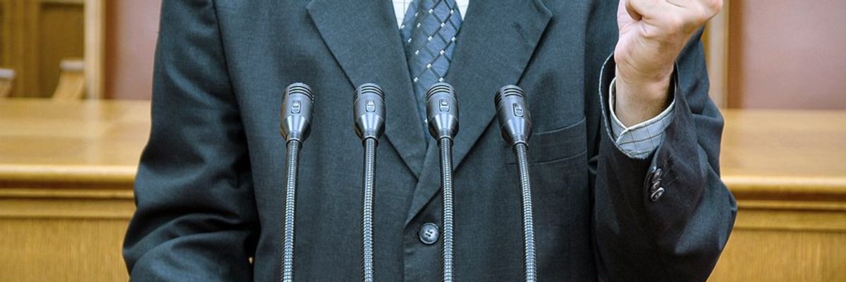 Torso of a politician in a suit standing at a podium shaking his finger.