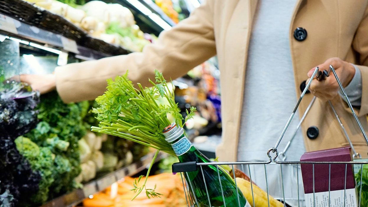 Torso of a woman holding a shopping basket while reaching for produce on a supermarket shelf