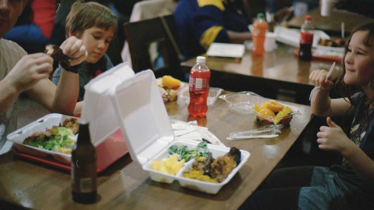 two children sitting at a cafeteria dining table eating from styrofoam and plastic containers.