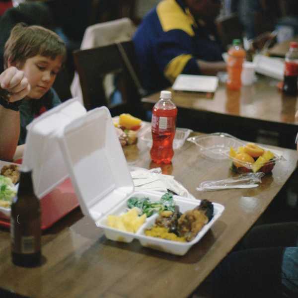 two children sitting at a cafeteria dining table eating from styrofoam and plastic containers.