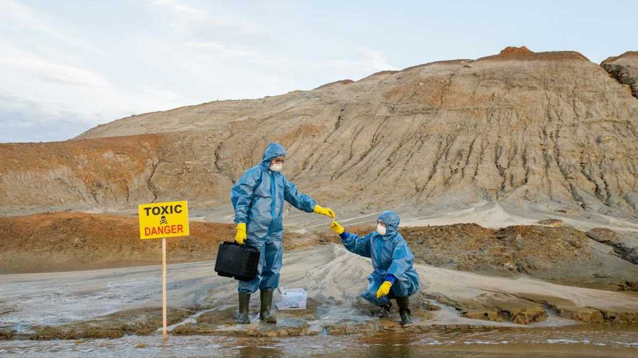 Two field workers in protective garb, sample water near a mining site adjacent to a sign reading Toxic Danger
