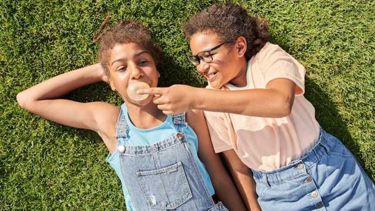 Two girls lying on the grass with one girl blowing a bubble with her chewing gum