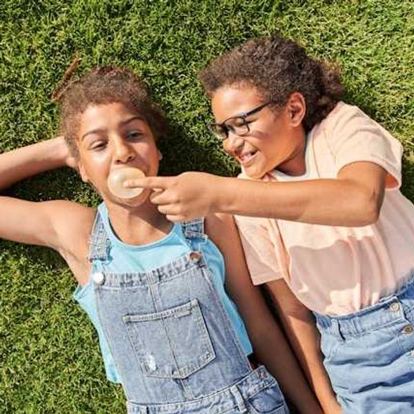Two girls lying on the grass with one girl blowing a bubble with her chewing gum
