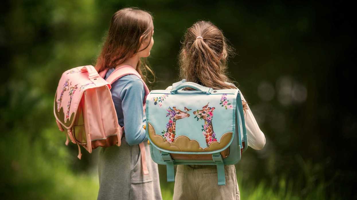 Two girls stand with their backpacks near water and trees.