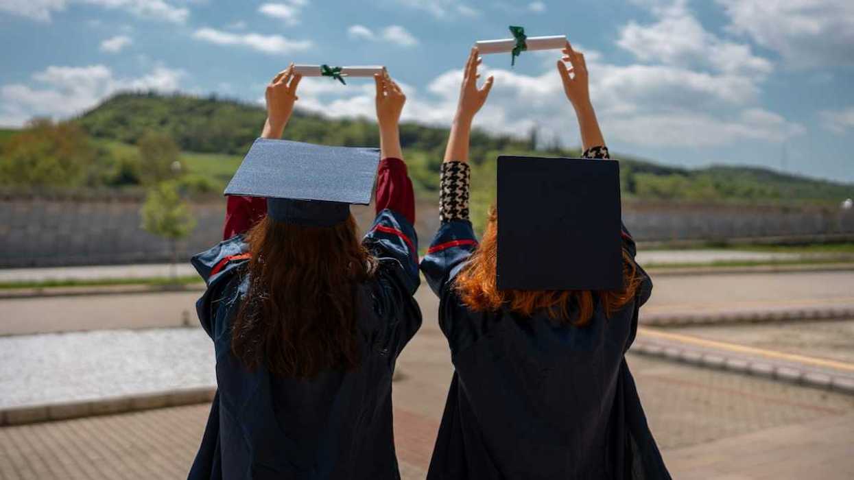 Two graduates with backs to camera holding their rolled-up diplomas above their heads, against the sky