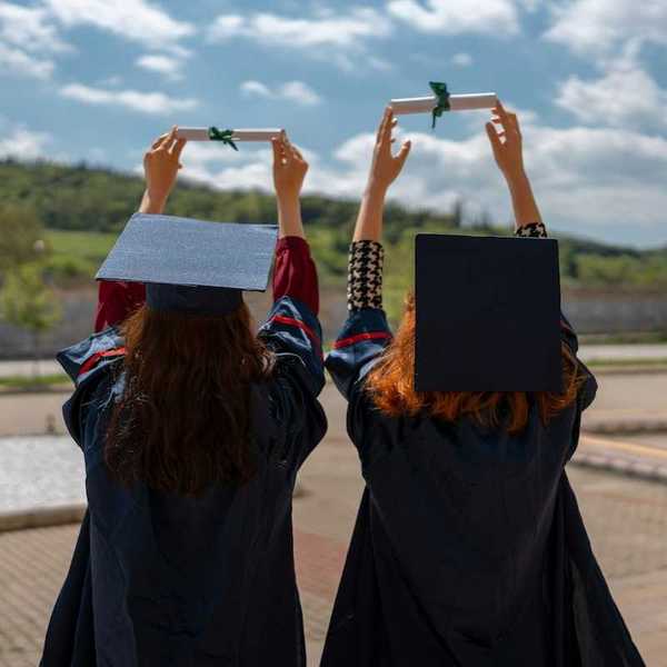 Two graduates with backs to camera holding their rolled-up diplomas above their heads, against the sky
