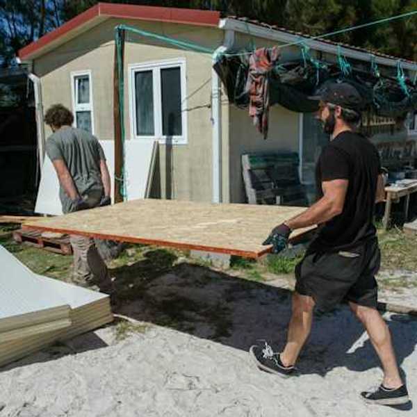 Two men carrying a plywood sheet past a manufactured home