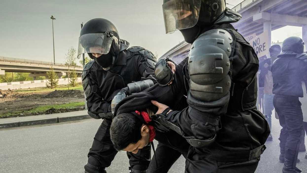 Two men dressed in protective gear - pads and helmets with face shields - while either detaining or helping a third man