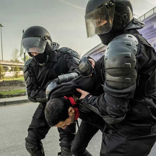 Two men dressed in protective gear - pads and helmets with face shields - while either detaining or helping a third man