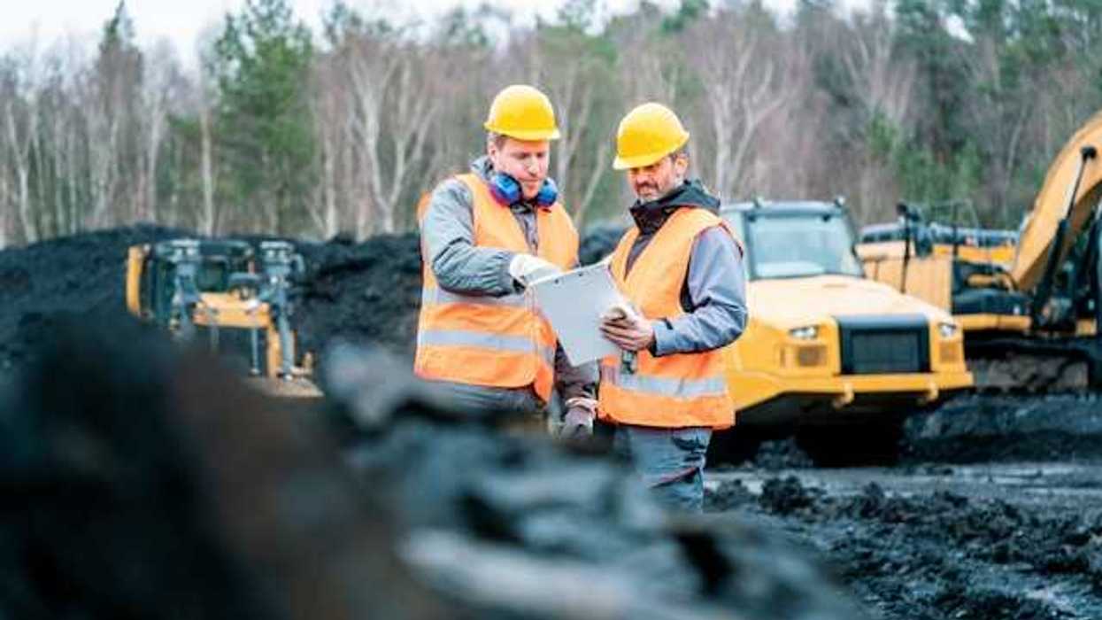 Two men wearing orange safety vests standing in front of piles of coal