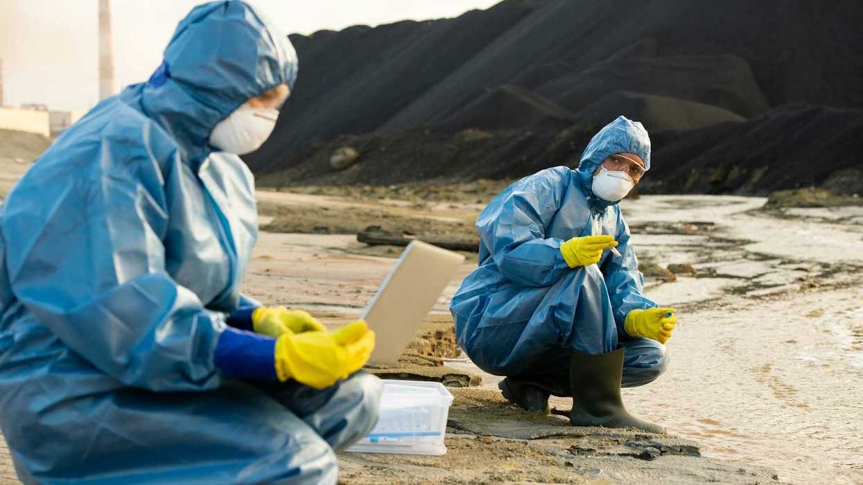 Two people clad in blue haz-mat suits sampling water running adjacent to a coal waste pile