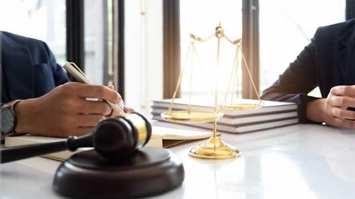 Two people sitting at a desk with a gavel, large legal books, and a set of scales
