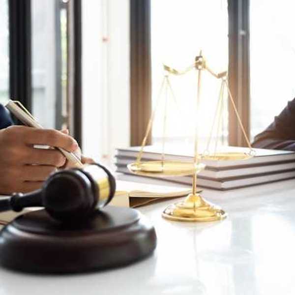 Two people sitting at a desk with a gavel, large legal books, and a set of scales