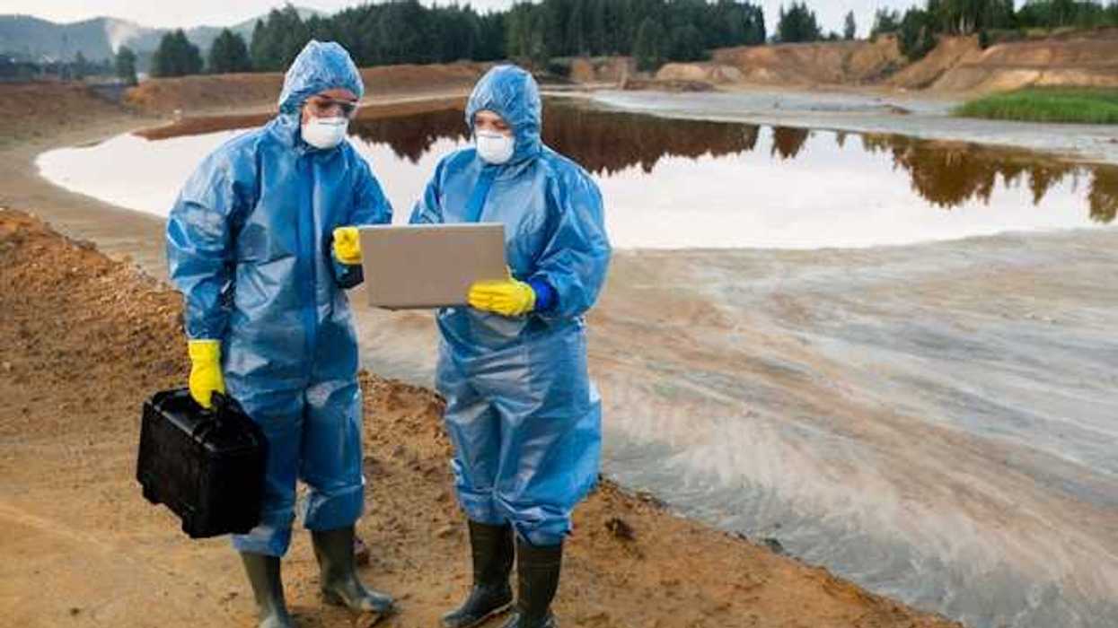 Two people wearing protective gear looking at a laptop in front of a mining waste pond