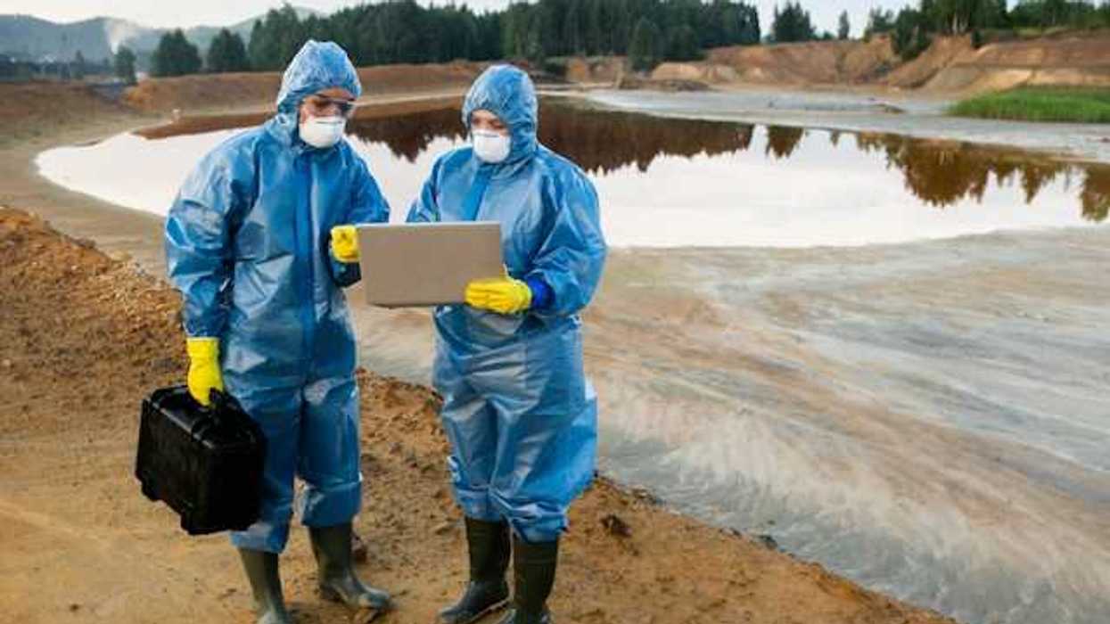 Two people wearing protective gear looking at a laptop while standing in front of a dirty pond