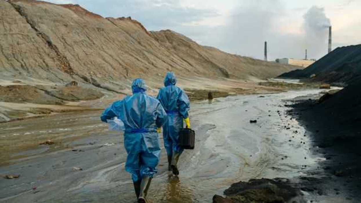 Two scientists in blue protective outfits walking along a stream near a large hill and industrial site in the distance