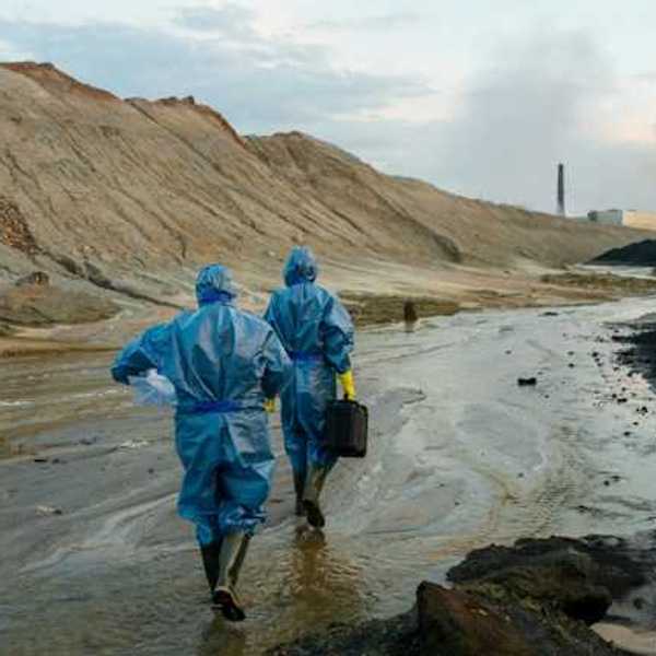 Two scientists in blue protective outfits walking along a stream near a large hill and industrial site in the distance