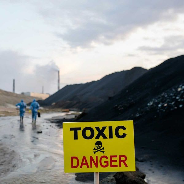 Two workers in blue hazmat suits walking towards coal-fired power plant with sign in foreground warning "toxic danger."