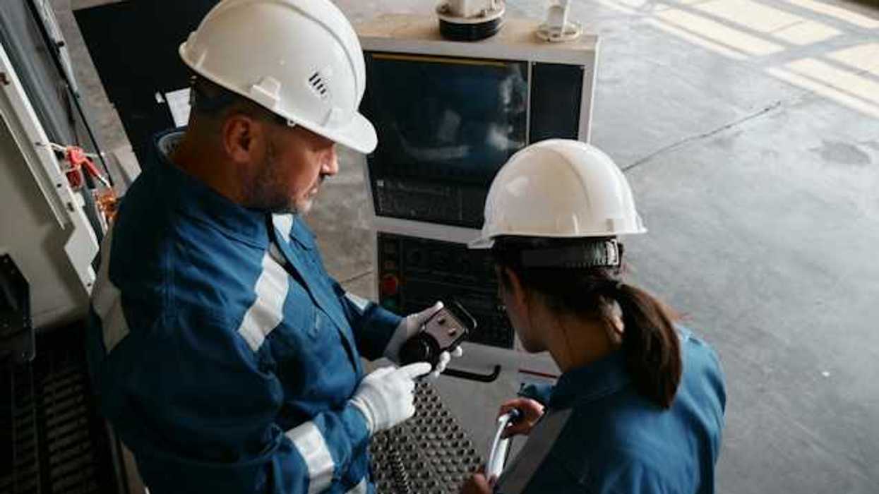 Two workers in blue jumpsuits and white hard hats looking at industrial equipment
