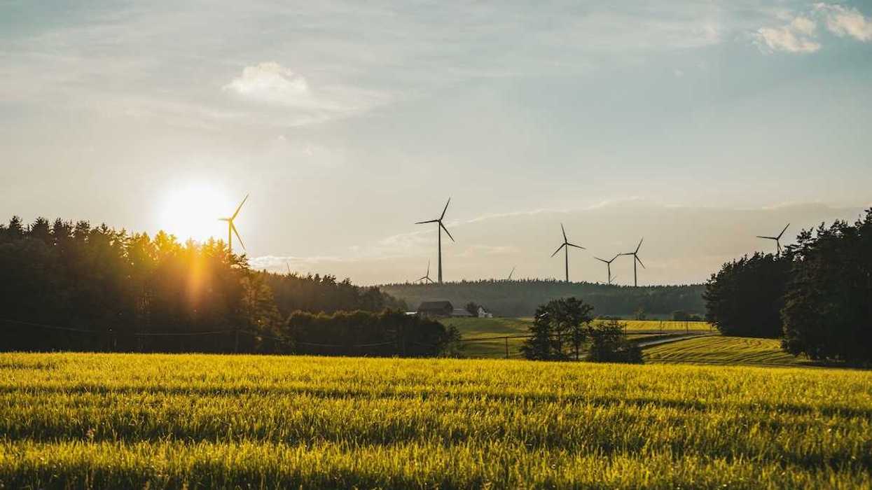 Wind turbines sited in a rural setting of forested hills with a farm and meadows in foreground