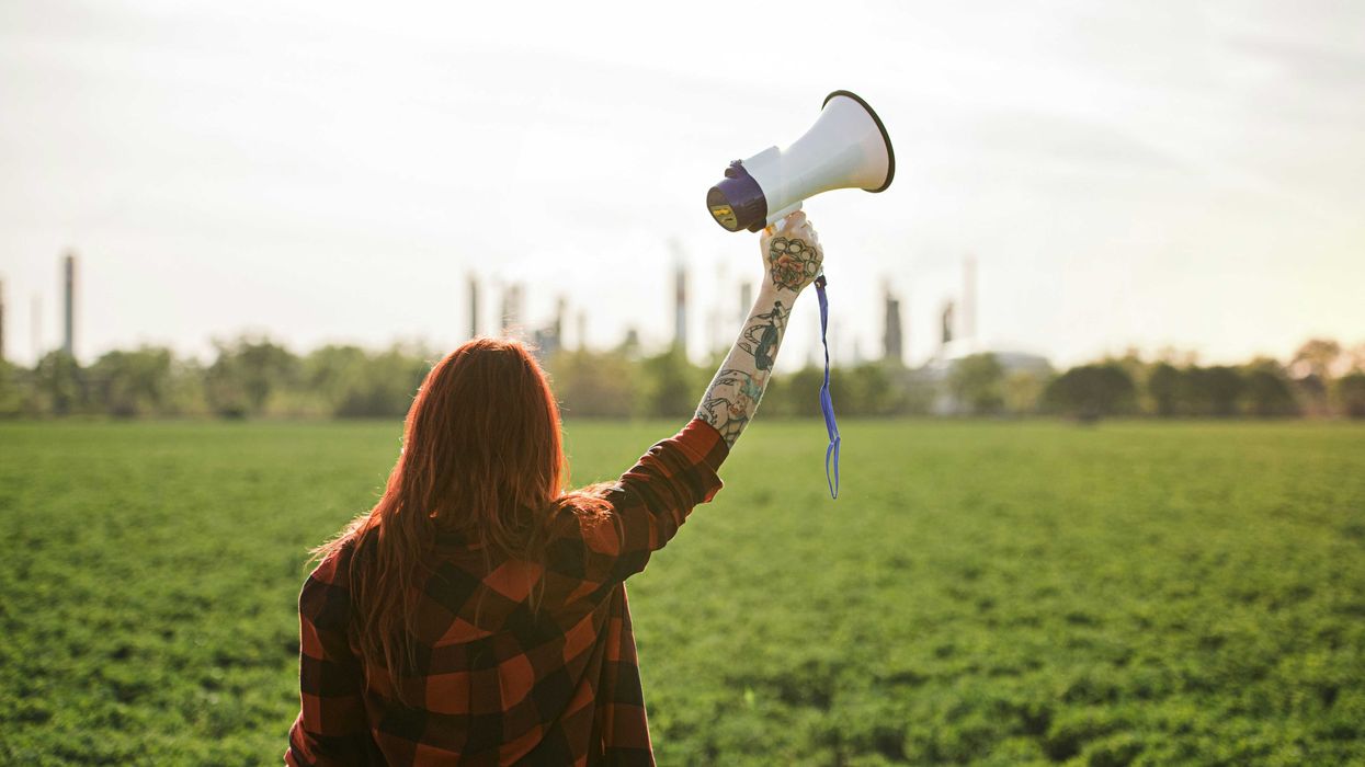 Woman holding up a megaphone facing a refinery across a green field.
