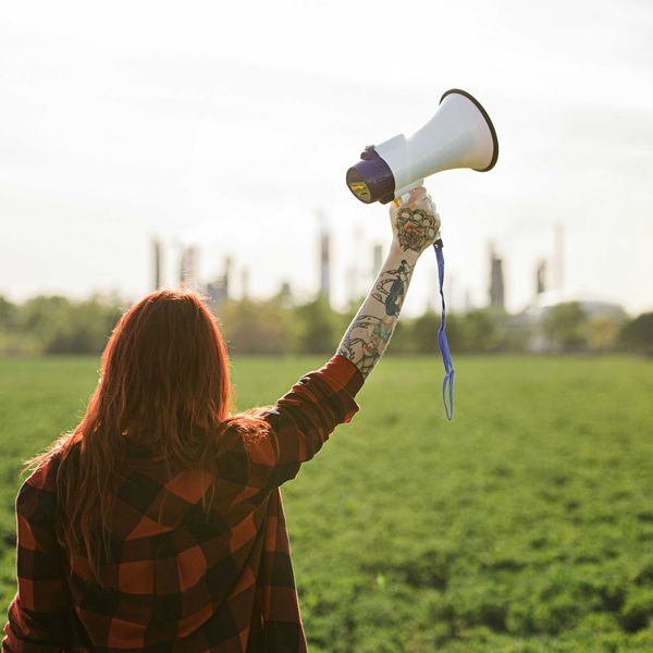 Woman holding up a megaphone facing a refinery across a green field.