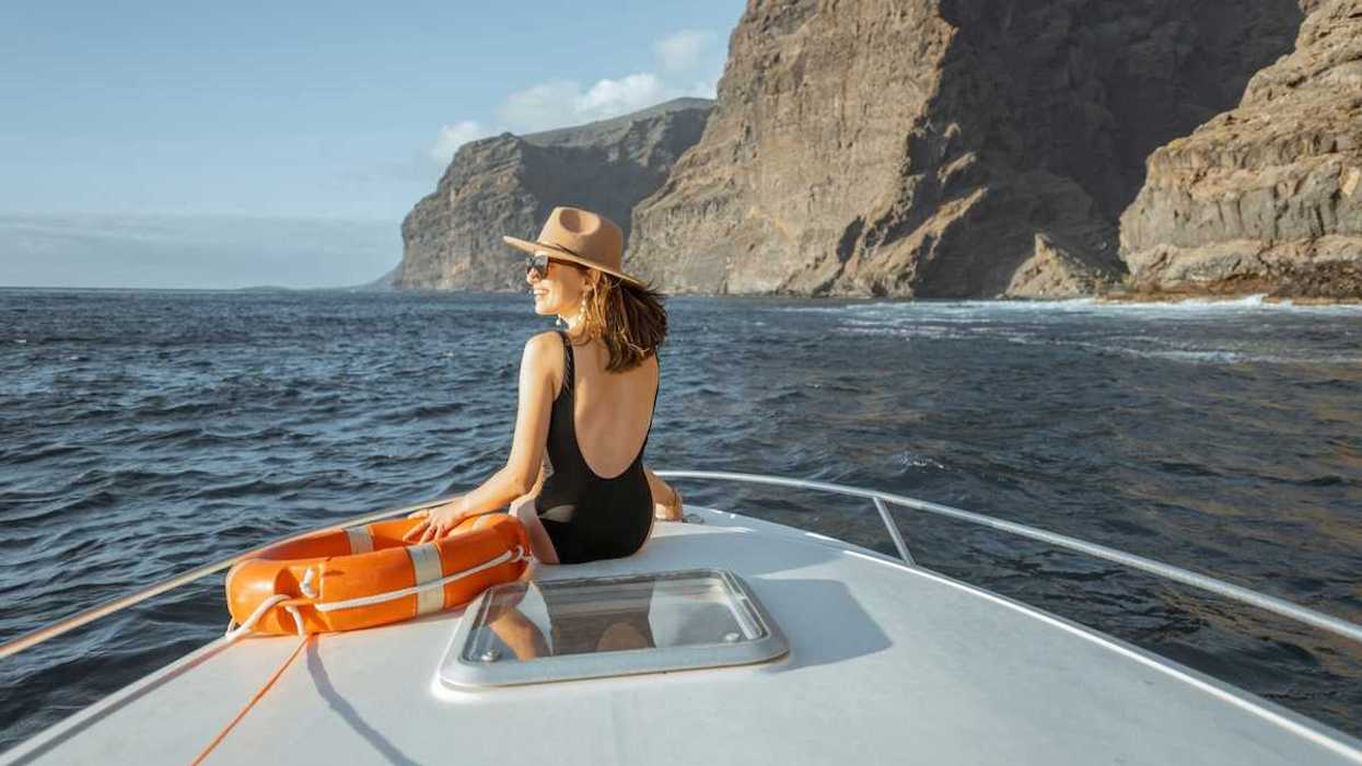 Woman in black bathing suit and broad brimmed hat next to an orange life preserver riding on the bow of a yacht