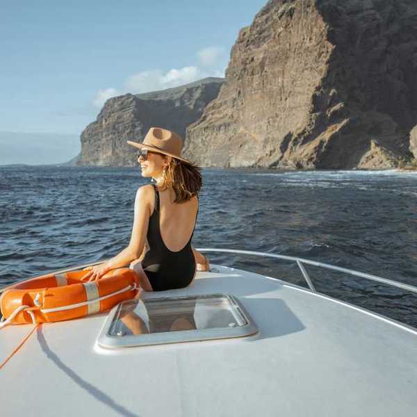 Woman in black bathing suit and broad brimmed hat next to an orange life preserver riding on the bow of a yacht