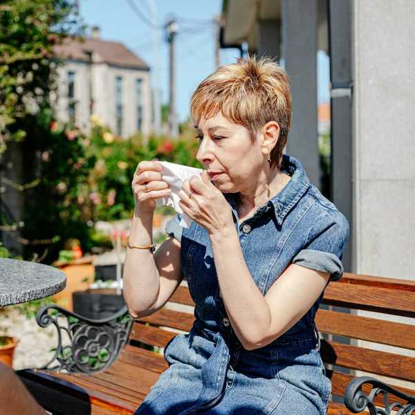 Woman in blue denim dress seated on an outdoor bench preparing to wipe nose with tissue