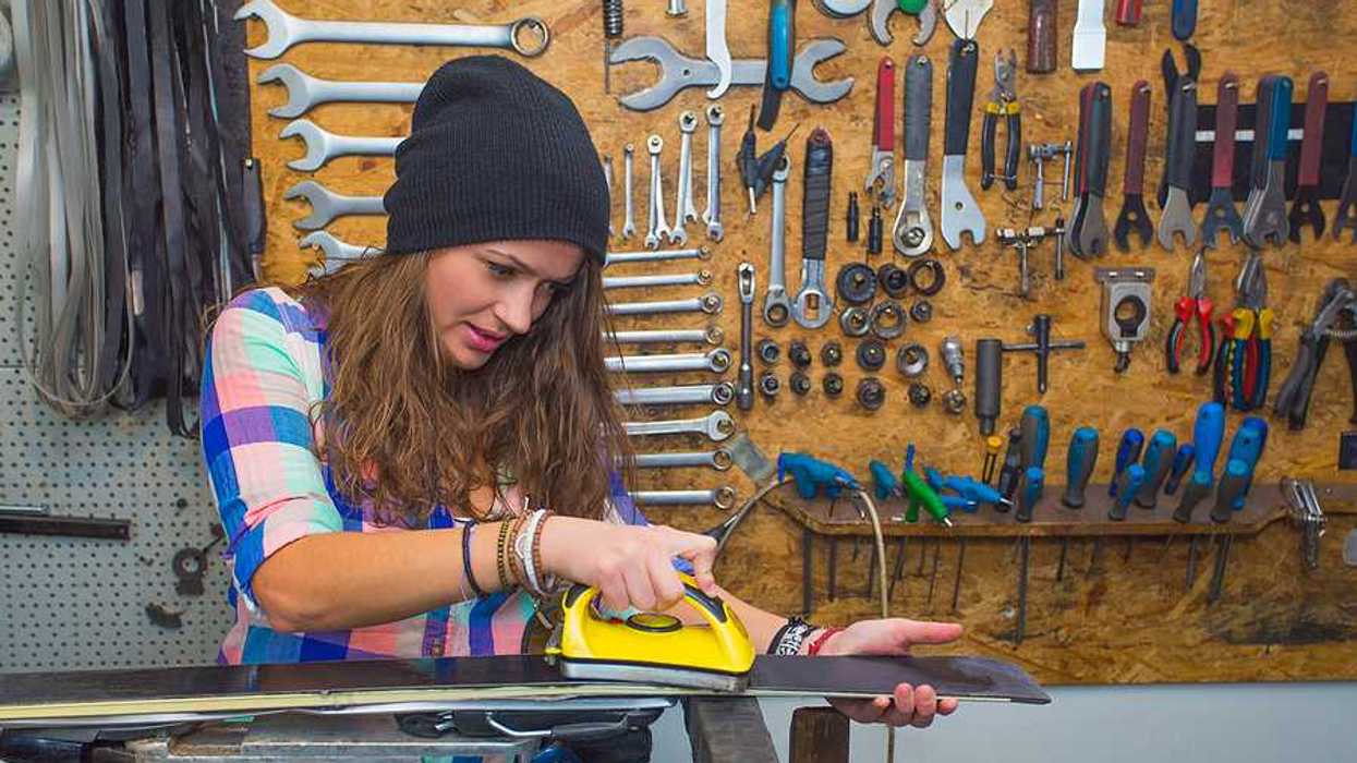 Woman in checkered shirt and hat hot-waxing a ski in a workshop.