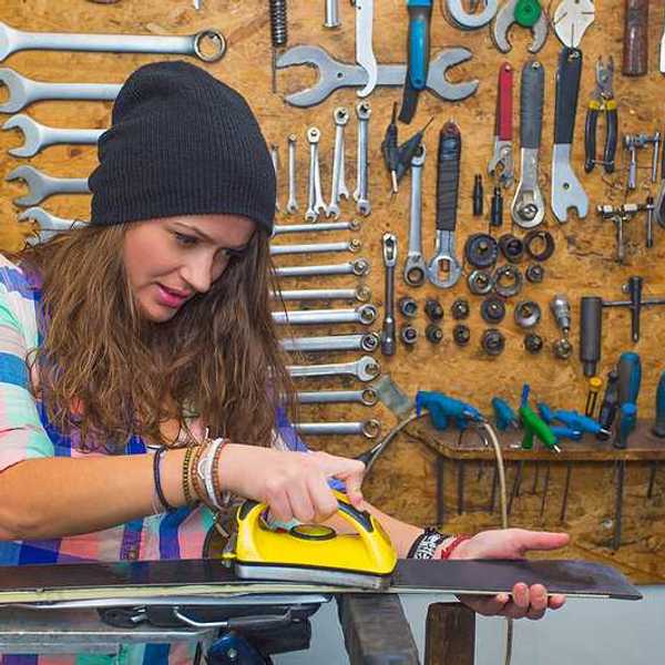 Woman in checkered shirt and hat hot-waxing a ski in a workshop.