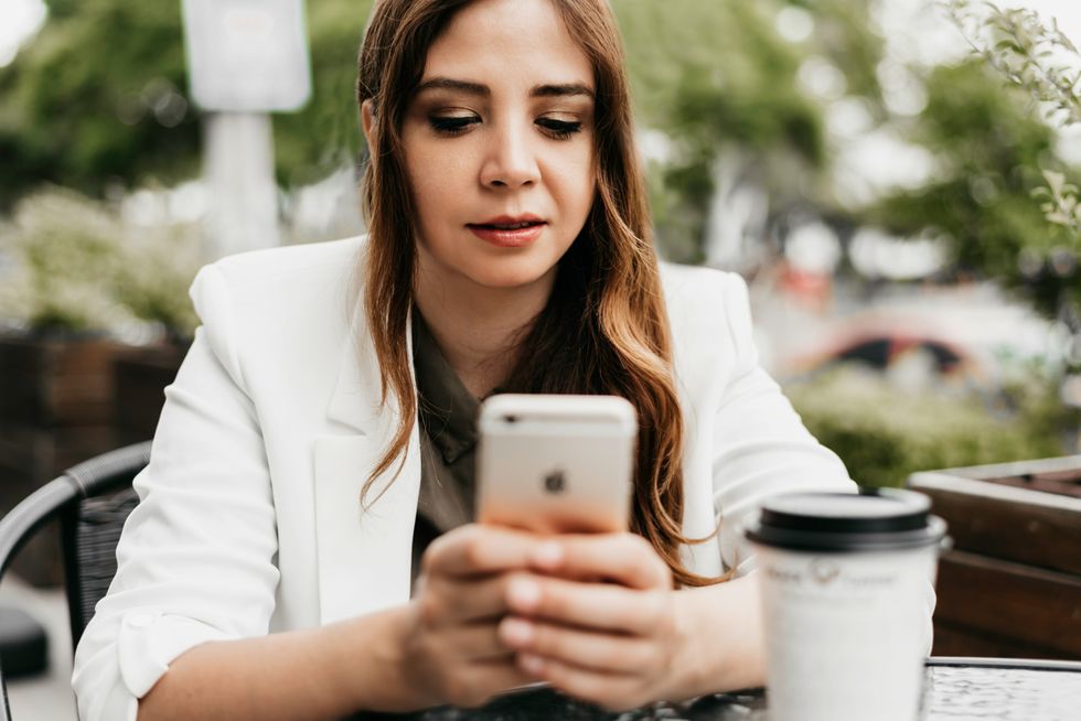 woman in white blazer seated outside holding white smartphone with a disposable mug of coffee in front of her.