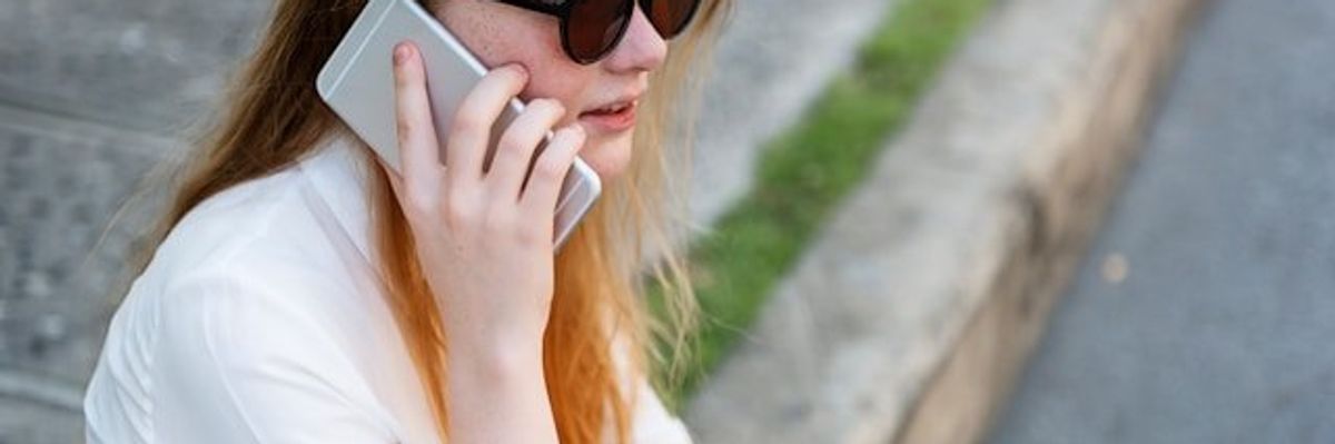 Woman in white shirt and sunglasses sitting on sidewalk listening to something on her phone.