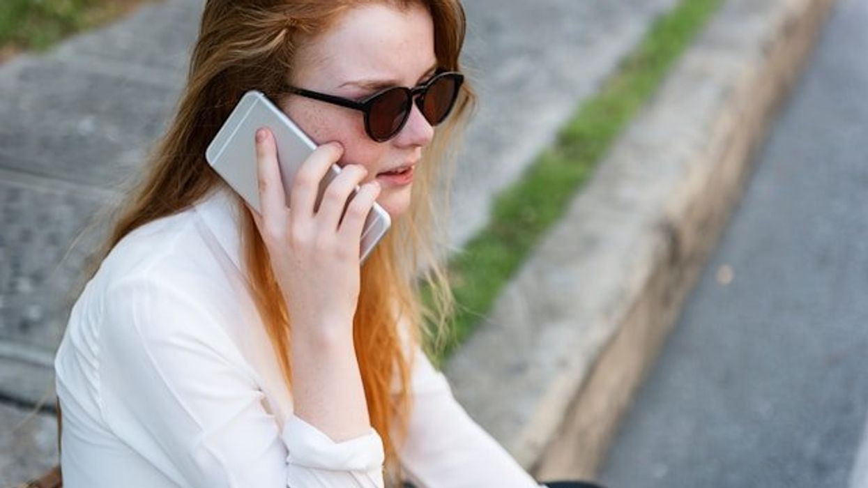 Woman in white shirt and sunglasses sitting on sidewalk listening to something on her phone.