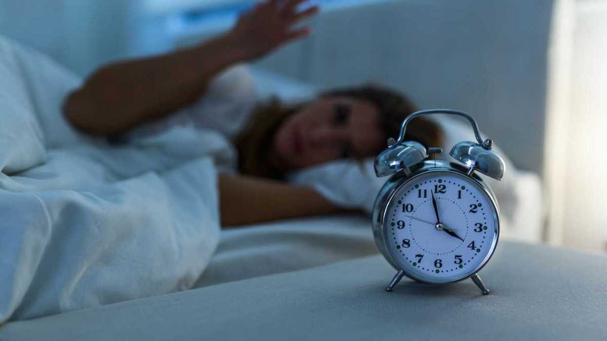 Woman lying awake in bed while an alarm clock indicates 3:58, presumably AM, exemplifies poor sleep.