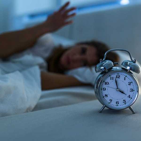 Woman lying awake in bed while an alarm clock indicates 3:58, presumably AM, exemplifies poor sleep.