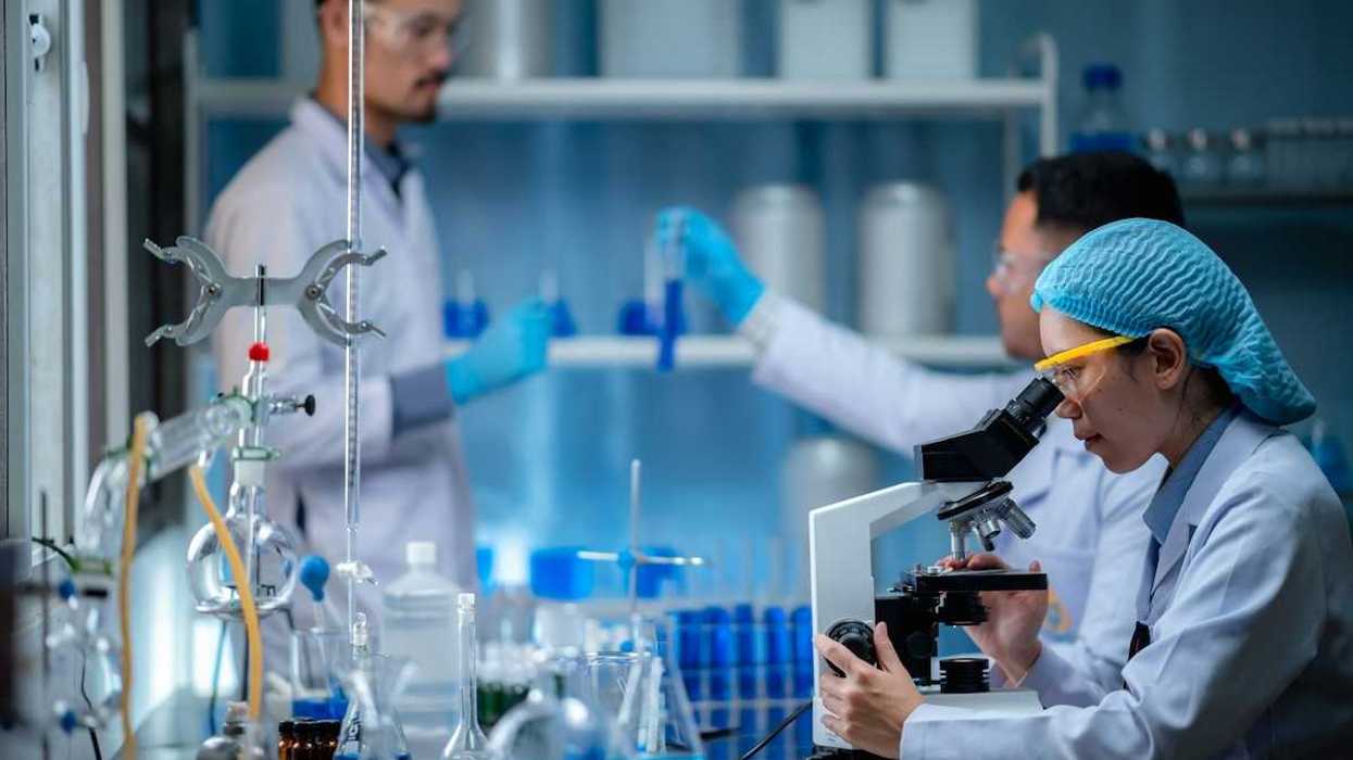 Woman peering into microscope while two men in lab coats and wearing blue gloves handle test tubes with a blue liquid