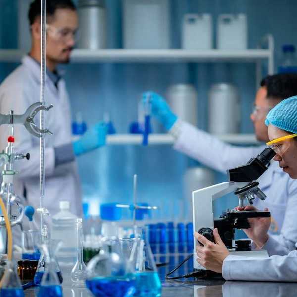 Woman peering into microscope while two men in lab coats and wearing blue gloves handle test tubes with a blue liquid