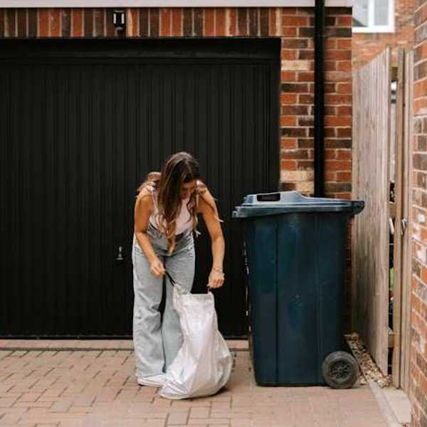 Woman placing a plastic trash bag on the grown next to a trash bin