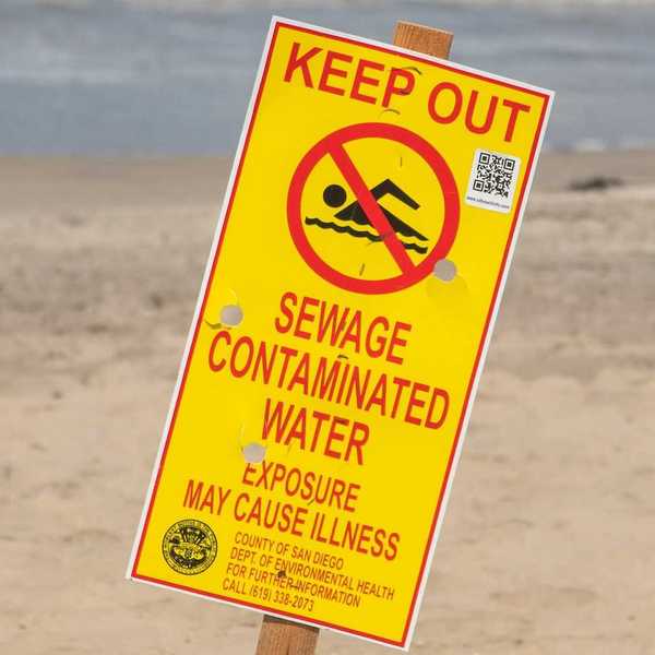 Yellow sign with red lettering reads "KEEP OUT, SEWAGE CONTAMINATED WATER. EXPOSURE MAY CAUSE ILLNESS"