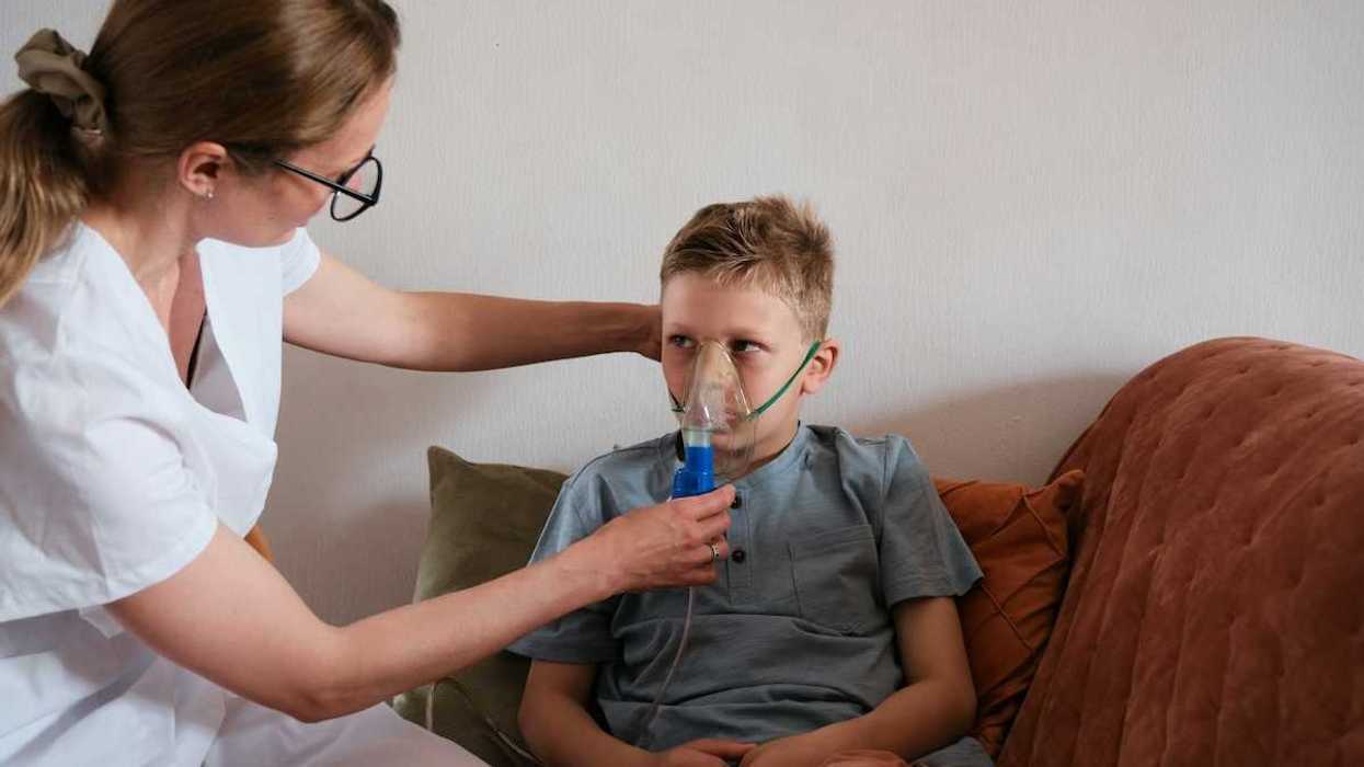 Young boy seated on sofa fitted with an inhaler being adfministered by a nurse in white