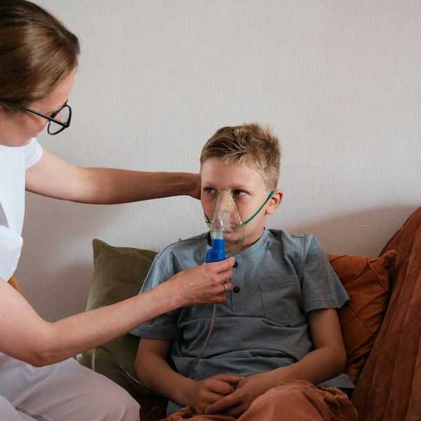 Young boy seated on sofa fitted with an inhaler being adfministered by a nurse in white