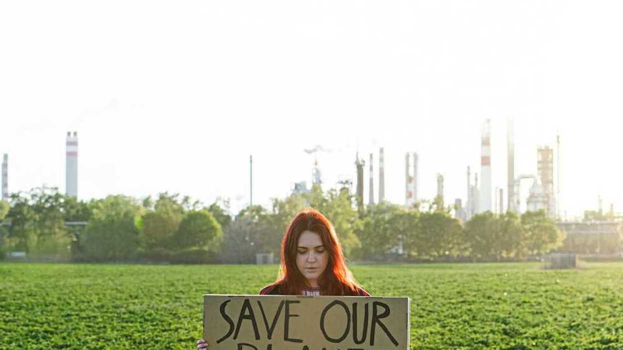 Young woman with polluting power plant in background holds a sign: "SAVE OUR PLANET"